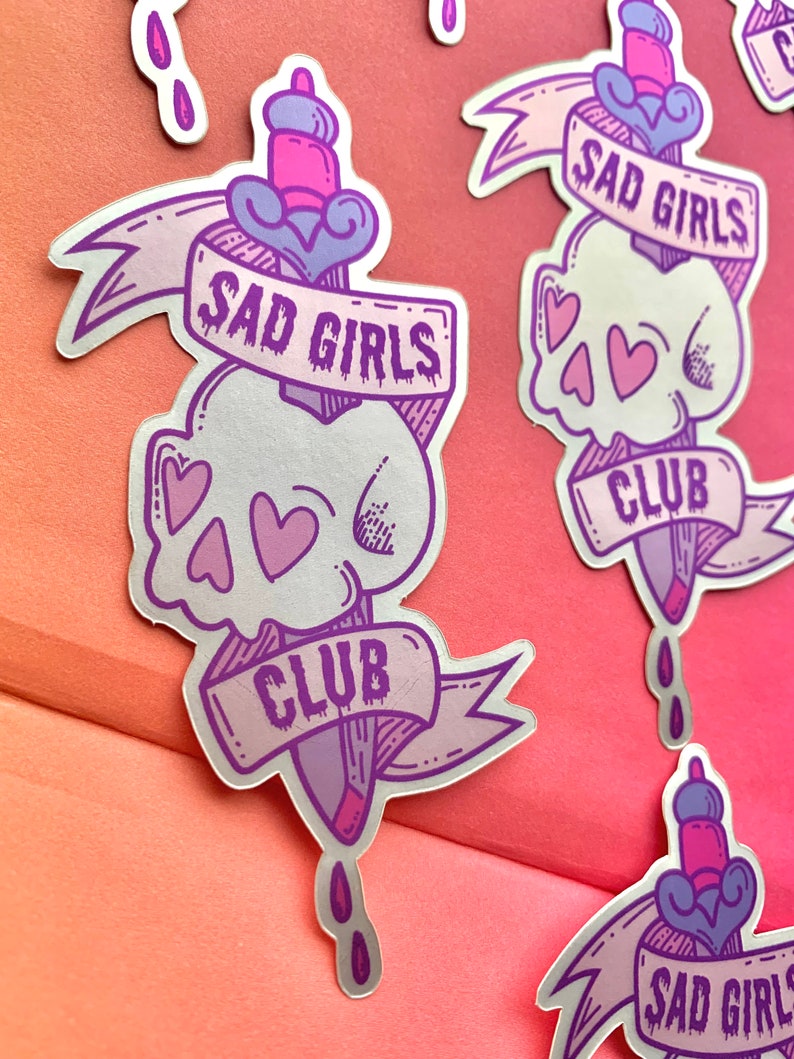 May include: A silver sticker with a purple and pink design. The sticker features a skull with hearts, a dagger, and a banner that reads "Sad Girls Club".
