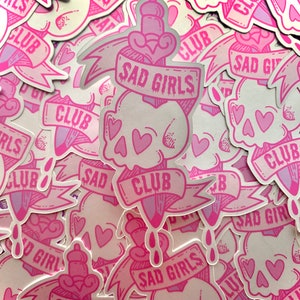 May include: A stack of pink and white stickers with a skull and dagger design. The stickers say "Sad Girls Club" and have pink hearts on them.