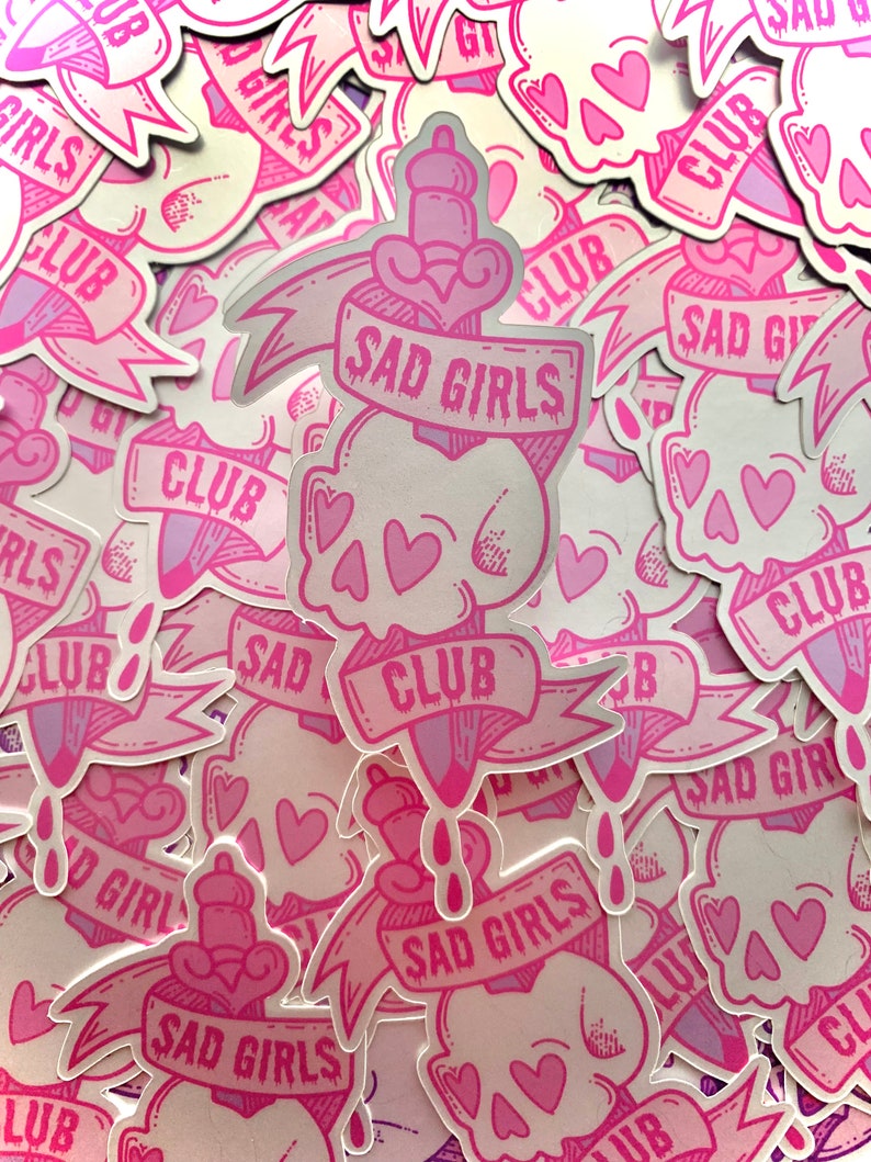 May include: A stack of pink and white stickers with a skull and dagger design. The stickers say "Sad Girls Club" and have pink hearts on them.