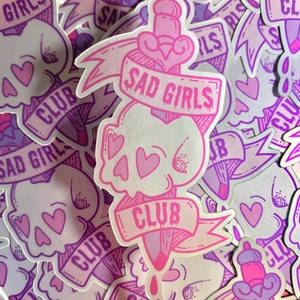 May include: A pink and purple sticker with a skull and dagger design. The sticker says "Sad Girls Club" in a pink banner.