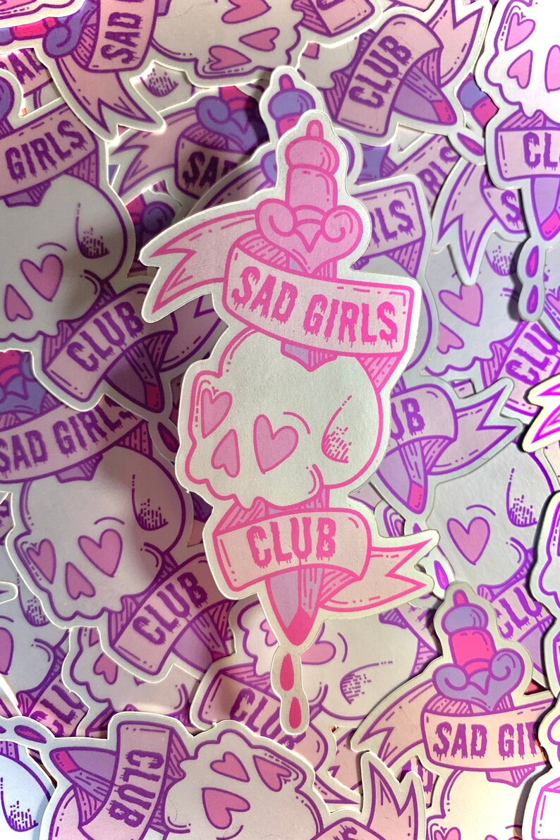 May include: A pink and purple sticker with a skull and dagger design. The sticker says "Sad Girls Club" in a pink banner.