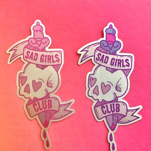 May include: Two silver holographic stickers with a skull and dagger design. The stickers feature the text "Sad Girls Club" in a purple font.