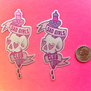 May include: Two silver holographic stickers with a skull and dagger design. The stickers feature the text "Sad Girls Club" in a pink and purple gradient.