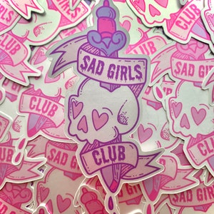 May include: A stack of stickers with a pink and purple design. The stickers feature a skull with a dagger through it, a banner that says "Sad Girls Club", and hearts.