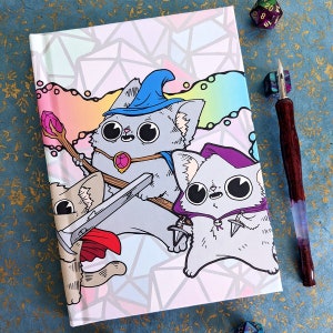 May include: A white notebook with a colorful geometric pattern featuring three cartoon cats wearing hats. One cat is wearing a blue wizard hat, another is wearing a purple hat, and the third is wearing a red hat. The notebook has a white background with a colorful geometric pattern.
