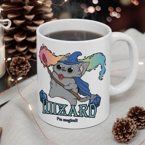 May include: White ceramic mug with a cartoon illustration of a gray cat wearing a blue wizard hat and holding a wand. The cat is surrounded by rainbow-colored swirls. The text "WIZARD I'm magical!" is printed on the mug.