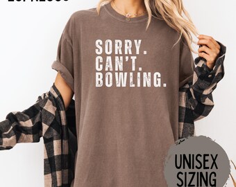 May include: Espresso-colored t-shirt with the text "SORRY. CAN'T. BOWLING." in white. The shirt has rolled-up sleeves and a relaxed fit. The words "UNISEX SIZING" are printed on a gray circle.