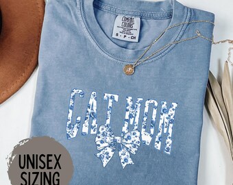 May include: A blue jean t-shirt with the text "Cat Mom" in white with a floral pattern. The text is in a large, bold font. A floral bow is below the text. The shirt is a unisex size.