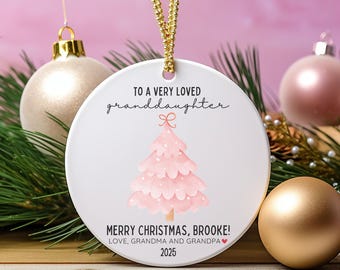 May include: A white ceramic Christmas ornament with a pink Christmas tree design and gold hanging cord. Text reads "TO A VERY LOVED granddaughter" and "MERRY CHRISTMAS, BROOKE! LOVE, GRANDMA AND GRANDPA 2025".