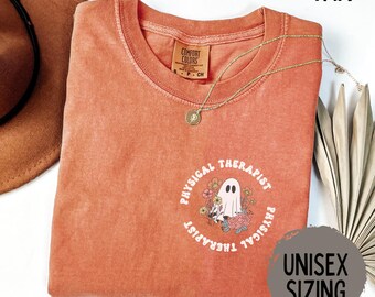 May include: A burnt orange t-shirt with a round graphic that says "Physical Therapist" around a ghost illustration. The shirt has a crew neck and short sleeves. A gold necklace with a pendant is on the shirt. The shirt is labeled "Unisex Sizing".