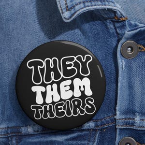 May include: A round, black button with the words "THEY THEM THEIRS" in white, stylized lettering. The button is set against a denim jacket background.