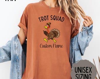 May include: A rust-colored t-shirt with the text "Trot Squad" above a cartoon turkey wearing a pilgrim hat. Below the turkey is the text "Custom Name". The shirt also has the text "Unisex Sizing" on a gray circle.