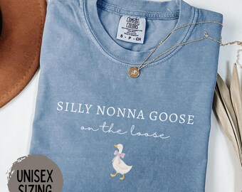 May include: Blue jean colored t-shirt with the text "SILLY NONNA GOOSE on the loose" and a cartoon goose with a pink bow. The shirt has a unisex sizing label and a gold necklace. The shirt is made of a soft material.