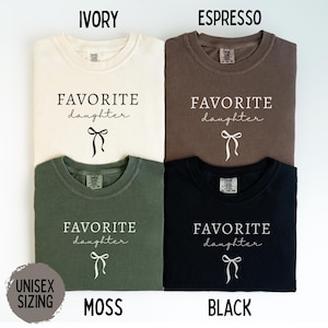 May include: Four unisex t-shirts in ivory, espresso, moss green, and black. Each shirt features the text "FAVORITE daughter" in a script font, with a bow graphic below. The shirts are labeled with their color names.