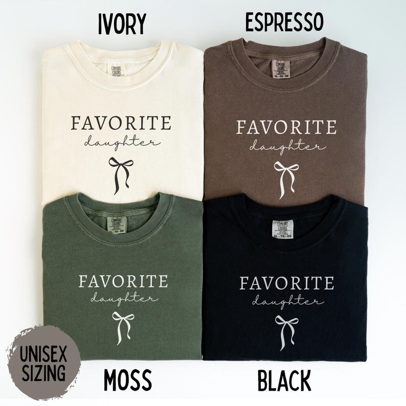 May include: Four unisex t-shirts in ivory, espresso, moss green, and black. Each shirt features the text "FAVORITE daughter" in a script font, with a bow graphic below. The shirts are labeled with their color names.