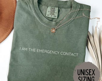 May include: A moss green t-shirt with the text "I AM THE EMERGENCY CONTACT" in white. A gold-colored necklace with a pendant rests on the shirt. The shirt has a crew neck and short sleeves. The tag reads "COMFORT COLORS".