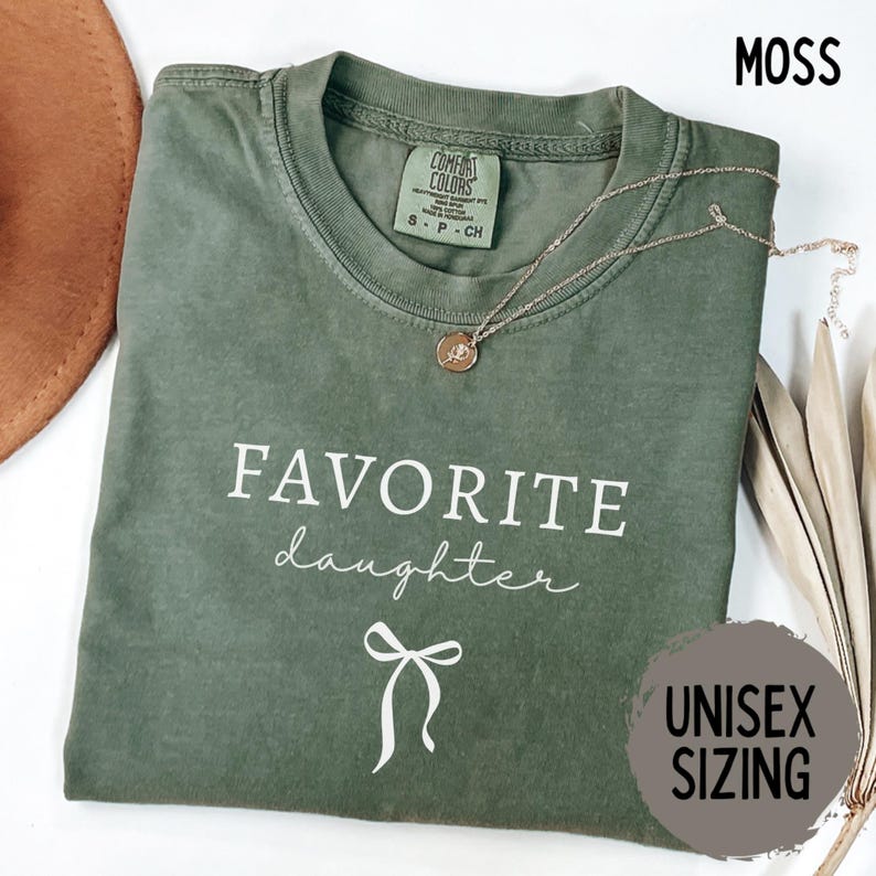 May include: A moss green unisex t-shirt with the words "FAVORITE daughter" in white script and a bow graphic. A gold necklace with a pendant rests on the shirt. The shirt is folded and has a tag that says "COMFORT COLORS".