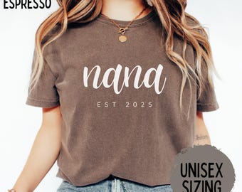 May include: Espresso-colored t-shirt with the word "nana" in white script, and "EST 2025" below. The shirt is labeled "UNISEX SIZING". The model is wearing a gold necklace and bracelet.