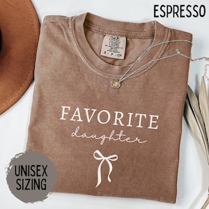 May include: Espresso-colored t-shirt with the words "FAVORITE daughter" in white script and a bow graphic. The shirt has a crew neck and short sleeves. A silver necklace with a pendant is draped across the shirt. The text "UNISEX SIZING" is also visible.