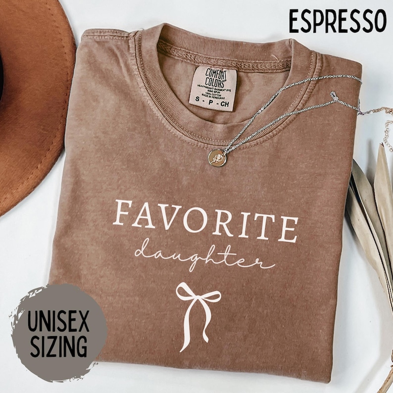 May include: Espresso-colored t-shirt with the words "FAVORITE daughter" in white script and a bow graphic. The shirt has a crew neck and short sleeves. A silver necklace with a pendant is draped across the shirt. The text "UNISEX SIZING" is also visible.