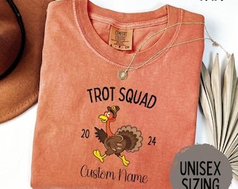 May include: A burnt orange t-shirt with the text "Trot Squad" and a cartoon turkey wearing a pilgrim hat. The numbers 20 and 24 are on either side of the turkey. The text "Custom Name" is below the turkey. The shirt is unisex sizing.