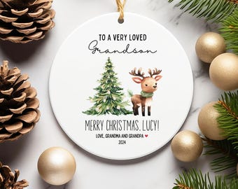 May include: White round Christmas ornament with a green Christmas tree and a brown reindeer with a red scarf. The ornament has the text "To a very loved Grandson Merry Christmas Lucy! Love, Grandma and Grandpa 2024" printed on it.
