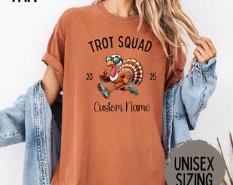 May include: A rust-colored t-shirt with the text "Trot Squad" above a cartoon turkey wearing sunglasses and running. The shirt also features the numbers "20" and "25" and the words "Custom Name". The shirt has rolled-up sleeves and a relaxed fit.