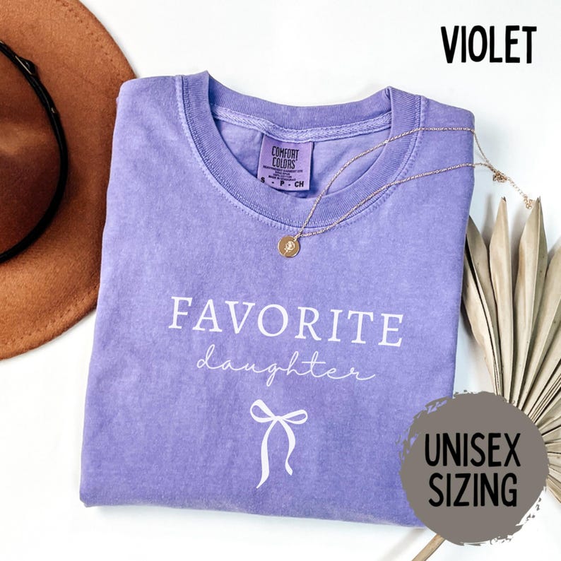 May include: A violet Comfort Colors t-shirt with the words "FAVORITE daughter" and a bow graphic. A gold necklace with a pendant rests on the shirt. The shirt is labeled "UNISEX SIZING".