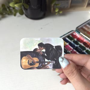 Może przedstawiać: Prostokątna naklejka z ilustracją przedstawiającą dwie całujące się osoby. Jedna osoba siedzi i gra na jasnobrązowej gitarze akustycznej, podczas gdy druga stoi i pochyla się w jej stronę. Scena utrzymana jest w delikatnej kolorystyce.