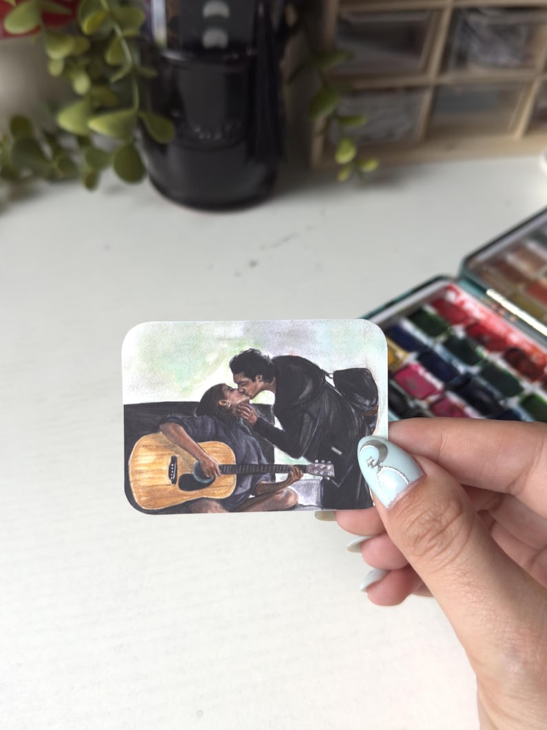 Może przedstawiać: Prostokątna naklejka z ilustracją przedstawiającą dwie całujące się osoby. Jedna osoba siedzi i gra na jasnobrązowej gitarze akustycznej, podczas gdy druga stoi i pochyla się w jej stronę. Scena utrzymana jest w delikatnej kolorystyce.