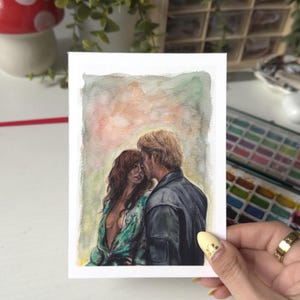 Op de afbeelding: Aquarel schilderij van een stel dat elkaar omhelst. De vrouw heeft lang, golvend roodbruin haar en draagt een groene top. De man heeft kort, blond haar en een donkere jas. De achtergrond heeft zachte aquarel wassingen.
