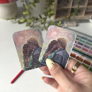 Könnte beinhalten: Zwei rechteckige Kunstdrucke mit einem sich umarmenden Paar, mit einer warmen Farbpalette. Das Kunstwerk zeigt einen Mann und eine Frau in einem zärtlichen Moment, mit einem sternenklaren Hintergrund. Die Drucke werden in einer Hand gehalten.
