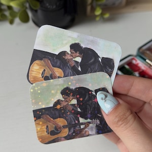 Könnte beinhalten: Zwei quadratische Aufkleber mit abgerundeten Ecken zeigen eine Aquarellillustration von zwei sich küssenden Personen, von denen eine eine Akustikgitarre hält. Ein Aufkleber hat einen holografischen Sterneffekt, der andere zeigt das Originalmotiv.