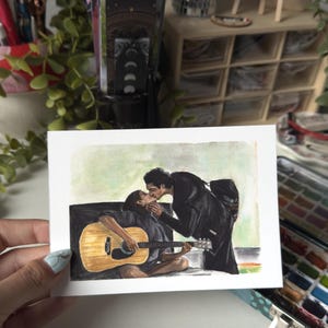 May include: A rectangular art print featuring a watercolour illustration of two individuals. One person sits while playing a light brown acoustic guitar, and the other person leans in to kiss them. Both figures are dressed in dark clothing against a light background.