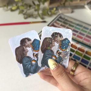 May include: Two rectangular stickers featuring a watercolor illustration of a couple embracing. The man wears a blue and white jersey with the number 44. The woman wears a blue jacket. The stickers have a glittery, sparkly effect.