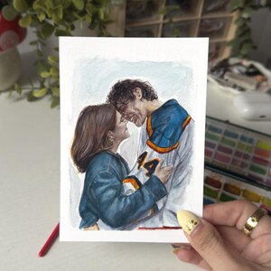 Könnte beinhalten: Aquarellmalerei eines sich umarmenden Paares, der Mann trägt ein blau-weißes Trikot mit der Nummer 44. Die Frau trägt eine blaue Jacke. Das Kunstwerk wird von einer Person mit einem goldenen Ring und gelbem Nagellack gehalten.