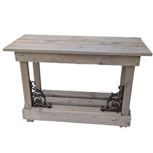 Pode incluir: Uma mesa de consola de madeira rústica com acabamento cinza desgastado. A mesa tem uma prateleira inferior e suportes de metal decorativos.