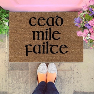 May include: A brown doormat with the text "cead mile failte" in black lettering. The doormat is on a stone surface with a pair of white shoes with black polka dots on top of it.