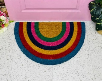 May include: A half-circle doormat with a rainbow design. The doormat is made of coir and has a natural brown color with stripes of red, orange, yellow, green, blue, and purple.