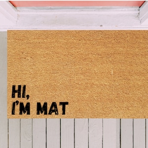Puede incluir: Una alfombra de coco marrón con el texto "Hi, I'm Mat" en letras negras.