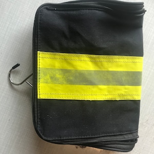Könnte beinhalten: Schwarze Reise-Kulturtasche mit einem leuchtend gelben Reflexstreifen. Die Tasche hat einen Reißverschluss und einen Metallhaken zum Aufhängen. Sie ist für die Aufbewahrung von Toilettenartikeln und persönlichen Gegenständen konzipiert.