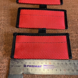 May include: Three red fabric rectangles with black trim and stitching, each with a black loop. A ruler shows measurements in inches and centimeters.