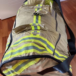 Puede incluir: Una bolsa de lona beige con múltiples bolsillos y rayas amarillas reflectantes. La bolsa tiene una correa negra para el hombro y un cierre de cremallera. La bolsa parece estar hecha de un material duradero, posiblemente resistente al fuego.