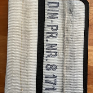 May include: A rectangular, light grey fabric item with a black border and a vertical black stripe. The text "DIN-PR.NR.81717" is printed vertically on the right side. The item appears to be a case or cover.