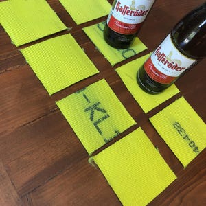 Könnte beinhalten: Zwei Flaschen Hasseröder Premium Pils Bier auf einem Holztisch mit gelben quadratischen Untersetzern. Die Flaschen sind teilweise von den Untersetzern verdeckt.