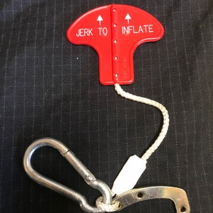 May include: Red plastic inflation device with a white rope and a silver carabiner clip. The device has two arrows pointing upwards and the text "JERK TO INFLATE" printed on it.