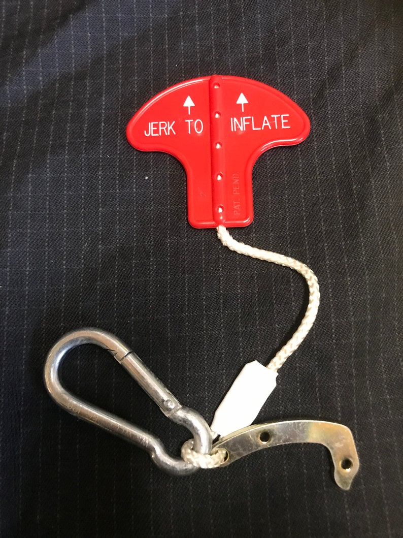 May include: Red plastic inflation device with a white rope and a silver carabiner clip. The device has two arrows pointing upwards and the text "JERK TO INFLATE" printed on it.