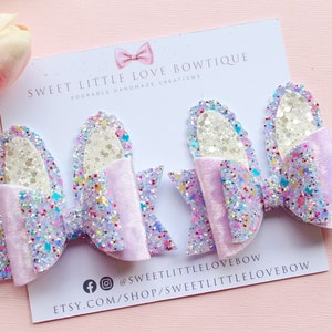May include: Three purple and white glitter hair bows with a light purple velvet centre. The bows are on a white card with the text "Sweet Little Love Bowtique Adorable Handmade Creations" and social media handles.