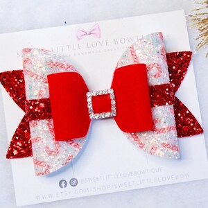 May include: A red and white glitter bow with a red velvet center and a rhinestone buckle. The bow is on a white card with the text "Sweet Little Love Bow Boutique" and a website address.
