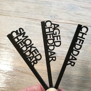 May include: Three black wooden cheese markers with the words "Aged Cheddar", "Smoked Cheddar", and "Cheddar" written on them.